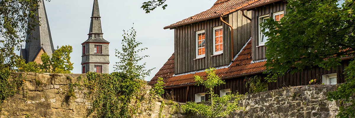 Kirchturmspitze und Turmspitze hinter einer Steinmauer, rechts ragt ein Hausdach über die Mauer hinaus